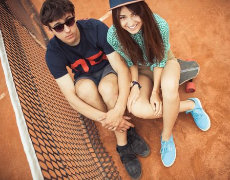 Young beautiful couple sitting on a skateboard on the tennis court