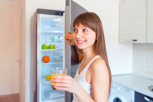 Young caucasian brunette girl with a glass of juice near the refrigerator