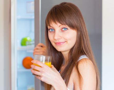 Young caucasian brunette girl with a glass of juice near the refrigerator