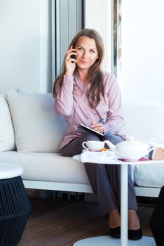 Happy businesswoman talking on the cellphone and writing in organizer in a coffee house