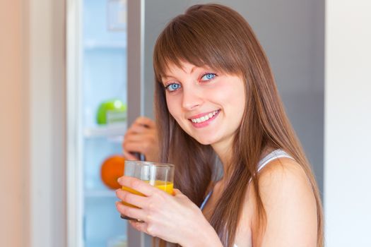 Young caucasian brunette girl with a glass of juice near the refrigerator