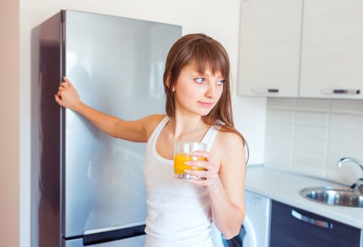 Young caucasian brunette girl with a glass of juice near the refrigerator