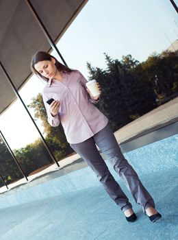 Young businesswoman with cellphone and cup of coffee while going against office building, outdoor