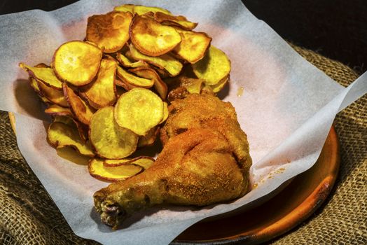 Homemade sweet potatoes chips and a chicken leg in a rustic clay plate on wood table