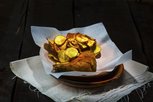 Homemade sweet potatoes chips and a chicken leg in a rustic clay plate on wood table