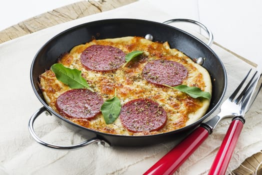 small salami pizza with basil leaf in a black pan and red fork and knife
