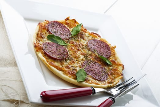 diagonal view of a small salami pizza on white plate with green basil leaf and red fork and knife on the right site