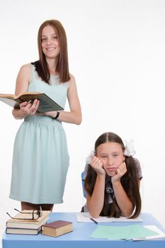 Teacher standing at desk behind which sits a tired student