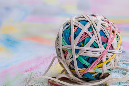 A ball of elastic bands of many different colours placed on a piece of paper that has also been coloured in many different vibrant colours.