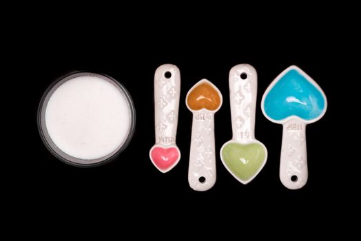 Heart shaped measuring spoons next to a bowl of sugar on an isolated black background.