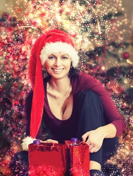 Beautiful middle age woman in red party santa hat sitting near the Christmas tree with gifts, with abstract background