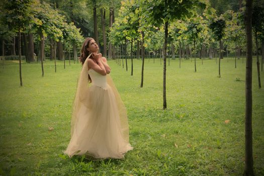 girl in white-golden gown in the summer park between young trees peers into sky, instagram image style