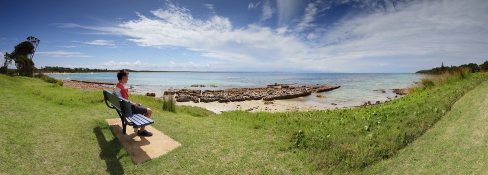 Grassy parklands right to the ocean at the southern end of Currarong Beach, Jervis Bay.  A tourist or visitor relaxes on a park bench soaking up the sun and taking in the views.   A shallow calm beach perfect for children at the sandy end, and they love exploring the rocks too.  9  vertical images stitched together, focus to foreground.