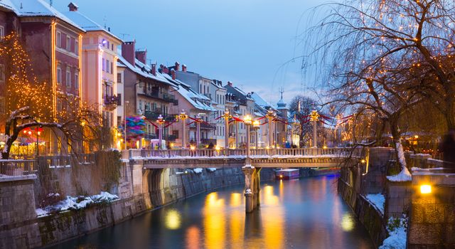 View of lively river Ljubljanica bank with Cobblers' Bridge in old city center decorated with Christmas lights. Ljubljana, Slovenia, Europe. Shot at dusk.