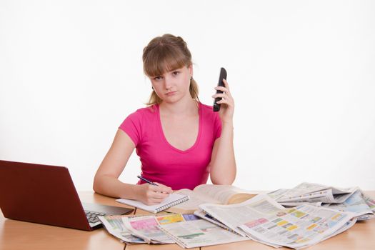 A young girl sits at a table with a pile of newspapers, encyclopedias and a laptop