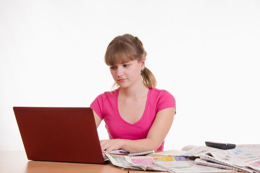 A young girl sits at a table with a pile of newspapers, encyclopedias and a laptop