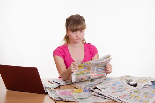 A young girl sits at a table with a pile of newspapers, encyclopedias and a laptop