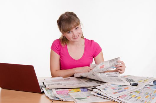 A young girl sits at a table with a pile of newspapers, encyclopedias and a laptop