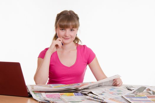 A young girl sits at a table with a pile of newspapers, encyclopedias and a laptop