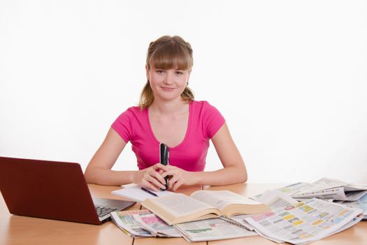 A young girl sits at a table with a pile of newspapers, encyclopedias and a laptop
