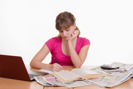 A young girl sits at a table with a pile of newspapers, encyclopedias and a laptop