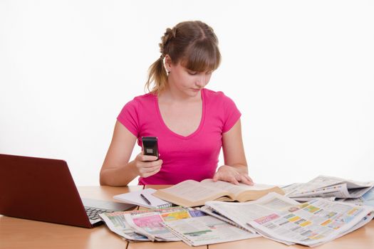 A young girl sits at a table with a pile of newspapers, encyclopedias and a laptop