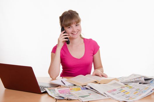 A young girl sits at a table with a pile of newspapers, encyclopedias and a laptop