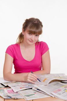 A young girl sits at a table with a pile of newspapers, encyclopedias and a laptop