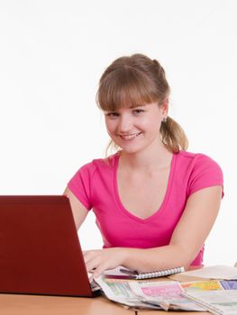 A young girl sits at a table with a pile of newspapers, encyclopedias and a laptop