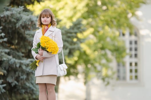 Young cute girl with a bouquet of yellow roses walks in autumn cool weather