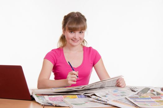 A young girl sits at a table with a pile of newspapers, encyclopedias and a laptop