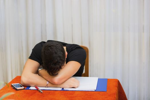 Tired, exhausted or despondent young man sitting at a table with a folder of paperwork leaning forwards with his head on his arms