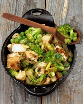Tasty Homemade Chicken Stew with Broccoli, Bell Pepper and Green Pea in Black Saucepan with Wooden Spoon isolated on Rustic Wooden background. Top View
