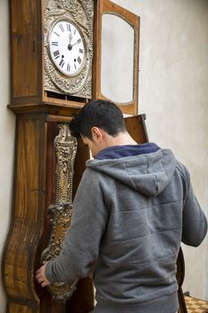 Young man checking the pendulum and winding the mechanism on a vintage long case clock standing with his back to the camera as he works
