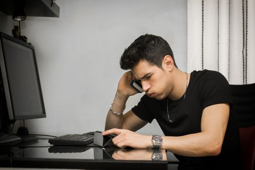 Young businessman taking a frustrating call on his mobile phone sitting grimacing as he works at his desktop computer in the office or at home