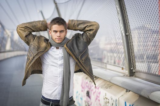 Handsome trendy young man standing on a sidewalk wearing a fashionable jacket and scarf with arms raised behind head looking at the camera