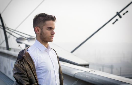 Handsome trendy young man standing on a sidewalk wearing a fashionable jacket and scarf in a relaxed confident pose looking away to a side