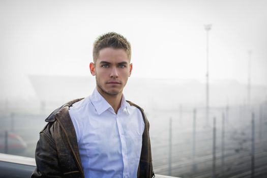 Handsome trendy young man standing on a sidewalk wearing a fashionable jacket and scarf in a relaxed confident pose looking at camera