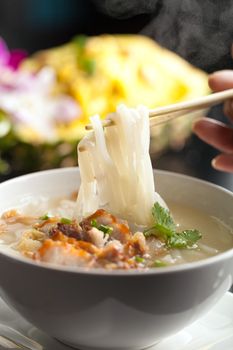 Closeup of a person eating Thai style crispy pork rice noodle soup from a bowl with chopsticks. Pineapple fried rice in the background.