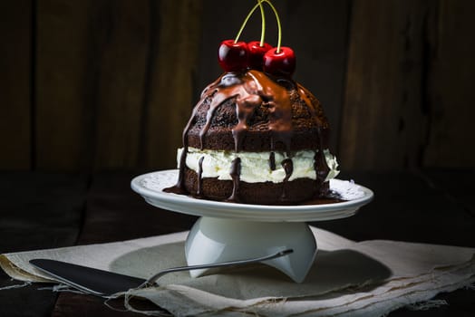 Delicious Chocolate Cake, Served on White Porcelain Tray, with Red Cherries on Top. Place on Top of Napkin at the Wooden Table with Spoon.