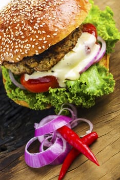 Close up Mouth Watering Hamburger with Lettuce, Tomatoes, Onions and Cream on Wooden Table with Onion Rings and Red Pepper.