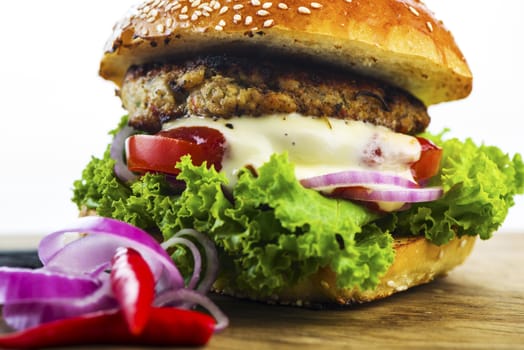 Close up Mouth Watering Hamburger with Lettuce, Tomatoes, Onions and Cream on Wooden Table with Onion Rings and Red Pepper.