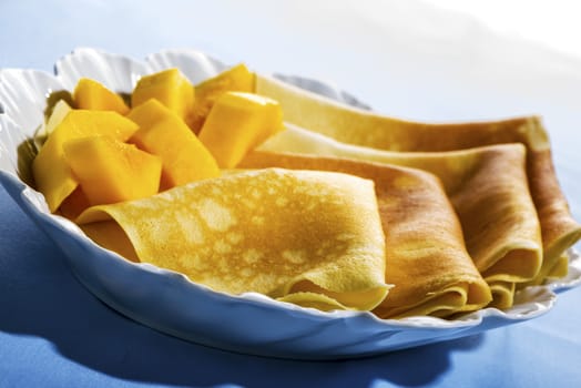 Freshly baked golden pancakes  or crepes neatly folded in a plate and served with fresh diced tropical fruit, close up view