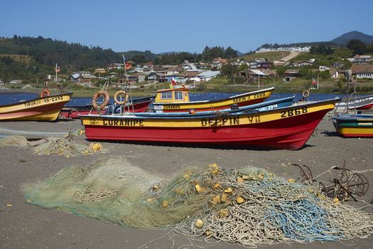 Colourfully painted wooden fishing boats drawn up on the on the beach in the small coastal town of Curanipe in Maule, Chile. The boats are pulled or pushed in and out of the sea using tractors.
