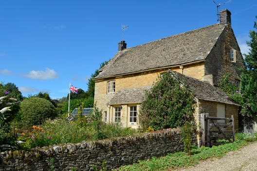 A honey-coloured Cotswold stone English cottage with a garden full of flowers and a British flag in Upper Slaughter, Cotswolds, UK.