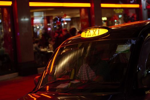 Low key detail of London black cab sign turned on at night, with red shop lights in the background.