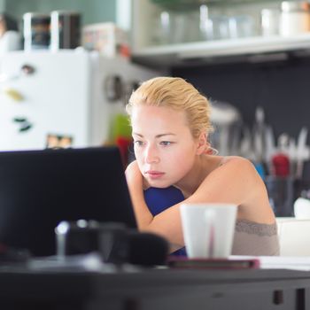 Female freelancer in her casual home clothing working remotly from her dining table in the morning. Home kitchen in the background.