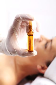 Gloved hand medical shows ampoule with a cosmetic preparation, in the background a woman waiting for surgery