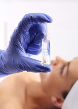 Gloved hand medical shows ampoule with a cosmetic preparation, in the background a woman waiting for surgery