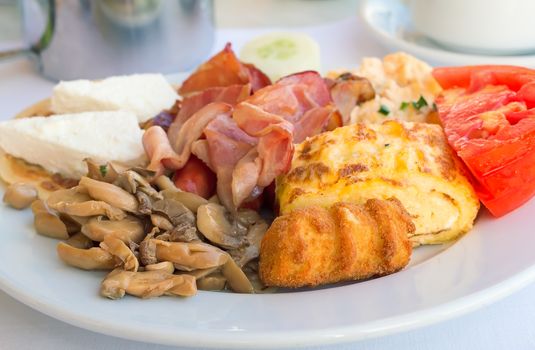 On the tablecloth table on a white plate are ham, scrambled eggs, cheese, mushrooms , tomatoes, varied and tasty food.